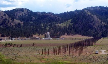 Photo of a series of posts creating a wall in the green plain, right of a white antenna surrounded by buildings with forested mountains in the background.