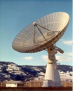 Photo of a fenced-in white parabolic antenna made of metal pointing towards the sky, located to the right of a small white building with snow-covered hills in the background