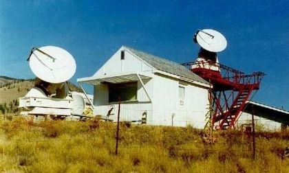 Photo of a rectangular white building and two antennas pointing towards the blue sky, surrounded by tall green grass.