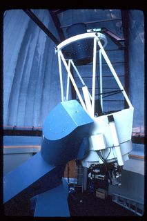 Photo of the blue and white structure of a telescope pointing towards the ceiling of a closed dome