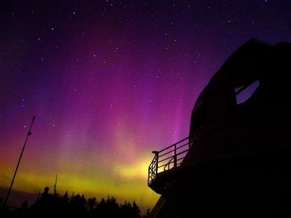 Backlit photo of a building with a dome facing yellow and purple Northern Lights under a star-filled sky