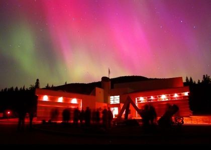 Night photo of a lit building with green and pink-tinged Northern Lights in the background. People are standing in front of the building looking skyward.