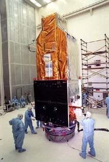 Photo of a yellow and silver-coloured telescope under construction in a white building. Men in protective blue clothing are working around the telescope.