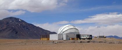 Photo of a white protective dome surrounded by rectangular buildings and a truck, taken in the desert with a grey mountain to the left and a cloudy blue sky as a backdrop.