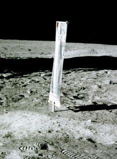 Photo of a vertical strip of cloth installed on the lunar soil behind traces of footprints on the ground with a very black sky in the background