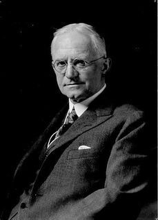 Black and white photo of a man wearing round eye glasses, a suit and tie, and looking at the lens